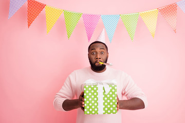 Photo of funny dark skin guy visit family meeting mother birthday noisemaker in mouth hold big giftbox wear sweater paper cap dotted flags hang pink color background