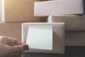 Fototapeta premium The small note attached on the product box wrapped by natural brown paper is captured or pulled by the hand of Asian woman. The stack of box are prepare to send or delivery product or gift to consumer