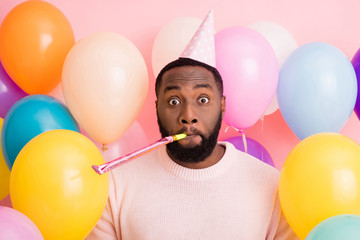 Photo of funky dark skin guy birthday surprise party surrounded many balloons blowing noisemaker crazy guest wear paper cone cap sweater on pink color background