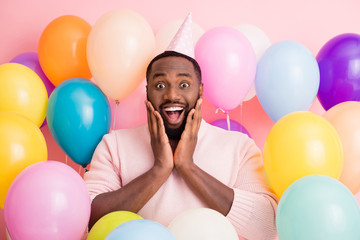 Photo of attractive dark skin guy amazed unexpected birthday surprise arms cheekbones surrounded many air balloons wear paper cone cap sweater on pink color background