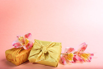 Trendy wrapped in golden textile in Furoshiki technique gift boxes and flowers Alstroemeria on pink. Valentine's day, mother's day, Women's day background. Zero waste holiday. Copy space.