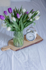 A bouquet of tulips in a glass vase, a white alarm clock on a wooden board stand on the bed. Spring.