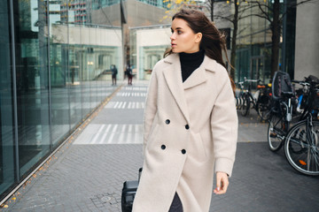 Young businesswoman in coat confidently walking around city street with suitcase