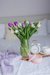 A bouquet of tulips in a glass vase, a white alarm clock, a candle, a note, a cappuccino mug on a wooden board are standing on the bed. Women's lace underwear. Breakfast in bed.