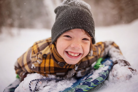 Close-up Portrait of a smiling boy in a woolly hat and duffel coat lying on a sledge in the winter snow, Wisconsin, USA