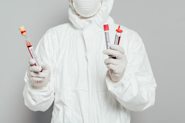 partial view of epidemiologist in hazmat suit holding test tubes with blood samples on grey background