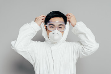 asian epidemiologist in hazmat suit and respirator mask putting hood on while looking at camera on...
