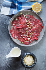 Round grey plate with carpaccio beef and condiments, vertical shot on a grey concrete background, flatlay