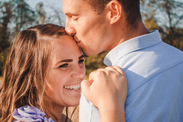 Fototapeta premium Close up of young couple in love hugging and smiling in hay field. Man hugs woman closely. Man kissing woman in head. Outdoor portrait of beautiful romantic couple in light clothes posing in summer
