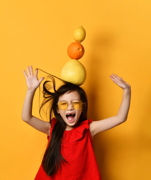 Little asian girl in sunglasses, red blouse, colored skirt. Screaming, holding pomelo, orange, lemon on her head, posing in studio