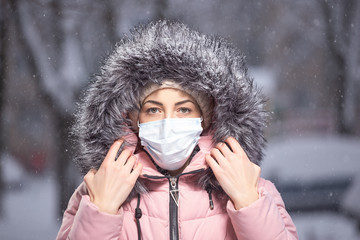 Portrait of woman in pink walking down the street in winter under snowfall in a protective mask to...