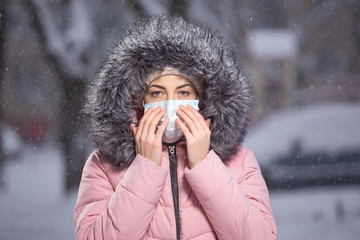 Portrait of woman in pink walking down the street in winter under snowfall in a protective mask to...