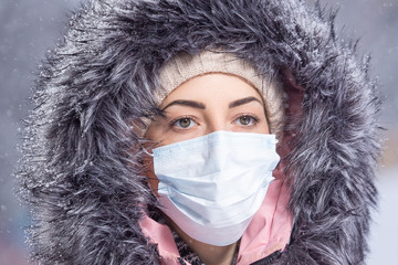 Portrait of woman in pink walking down the street in winter under snowfall in a protective mask to...