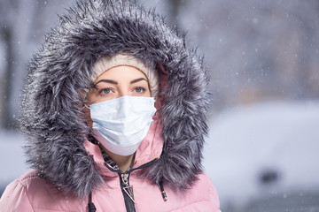 Portrait of woman in pink walking down the street in winter under snowfall in a protective mask to...
