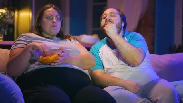 Overweight couple watching tv and eating fast food on couple