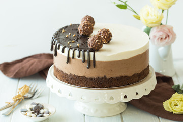 Whole mousse cake "three chocolate"  sprinkled with chocolate, and decorated candy. A plate with a cake stands on a white wooden table. © Maria