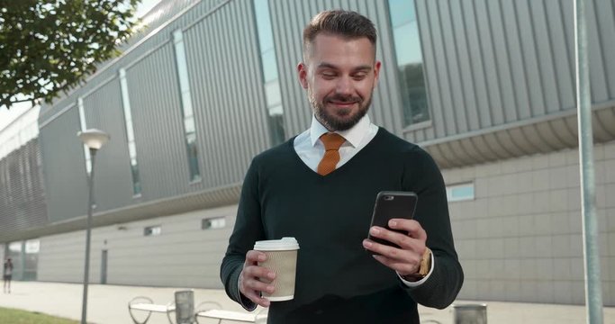 Attractive Office Worker Standing outdoor Office Building with Smartphone in his Hands. Having interesting conversation. Looking surprised at Screen while reading Messages. Having good Mood.