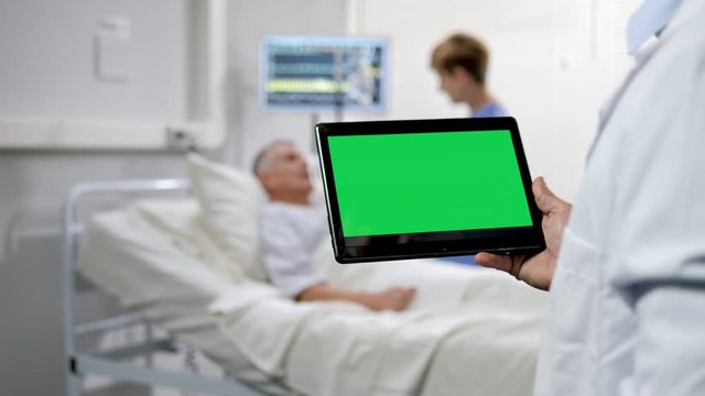 doctor holding a green screen tablet with a nurse taking care of a senior patient male lying in the hospital bed blurred in the background