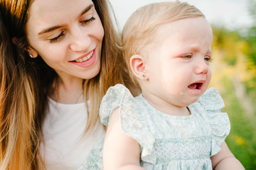Portrait of a little beautiful baby girl crying and mom on nature on summer day. The playing in the park with parents. Close Up. The concept of family holiday and time together.