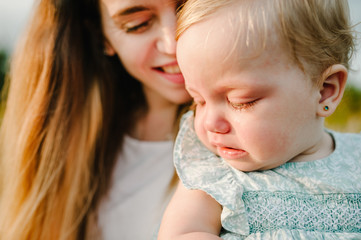 Portrait of a little beautiful baby girl crying and mom on nature on summer day. The playing in the park with parents. Close Up. The concept of family holiday and time together.