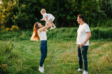 Naklejka na ściany i meble Portrait of a father, mother holds, throws up and spins the daughter on hands walking on nature on summer day vacation. Mom, dad and girl playing in the park. Concept of happy family.