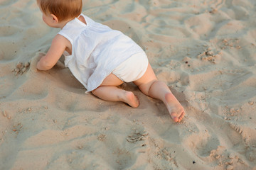 baby girl in white clothes and a straw hat sits on the white sand on the beach in summer - obrazy, fototapety, plakaty