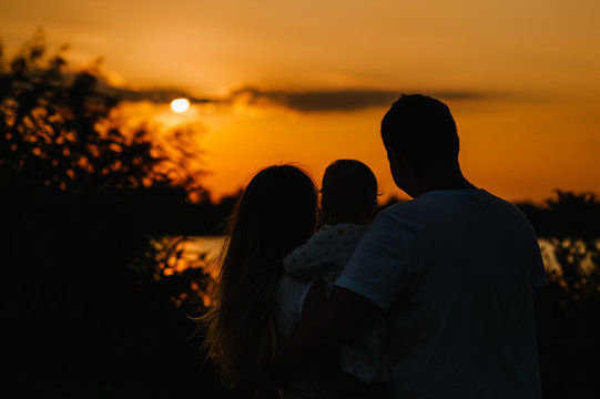 Mom, dad holds daughter stand back and looking on sunset. Portrait of a young family on nature, on vacation, outdoors.  The concept of family holiday.