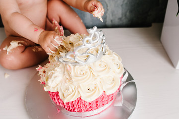 Eating cake hands. Portrait of caucasian baby girl celebrating her first birthday with gourmet white and pink cake with candle 1 year and crown on top of. cake smash first year concept.