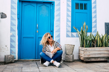 Beautiful adult blonde curly model woman sit down on the floor with paper book enjoying her alternative lifestyle - travel and influencer concept with blue house in background