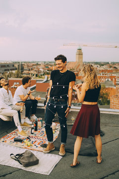 Playful woman tying string light around man while friends enjoying at rooftop party
