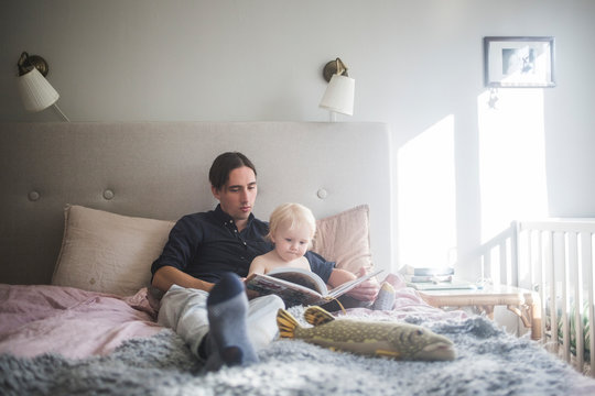 Father reading picture book with toddler son while sitting on bed at home