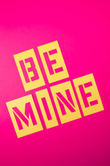 Be Mine Valentine's Day message spelled out in yellow stencil lettering on bright pink background