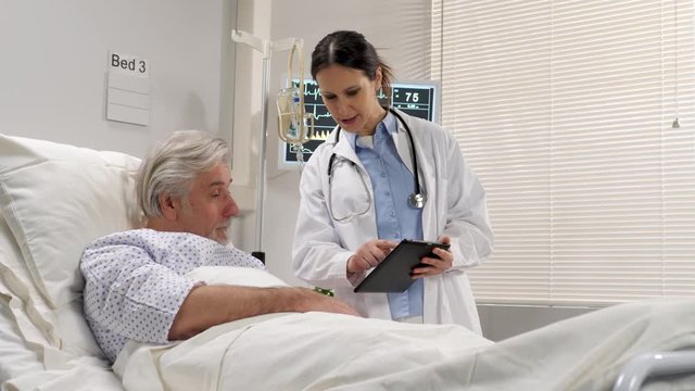 female doctor talking with a mature senior patient on the hospital bed showing to him medical results test on tablet comptur device
