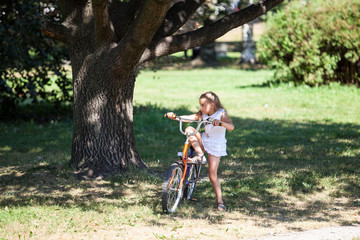 Fototapeta na wymiar Young girl trying to ride her bicycle in summer park on meadow with green grass