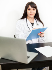 Portrait of young beautiful woman doctor or intern sitting at the desk with laptop and clipboard in hands, white background