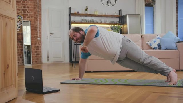 Overweight man doing exercise while watching tutorial on laptop at home