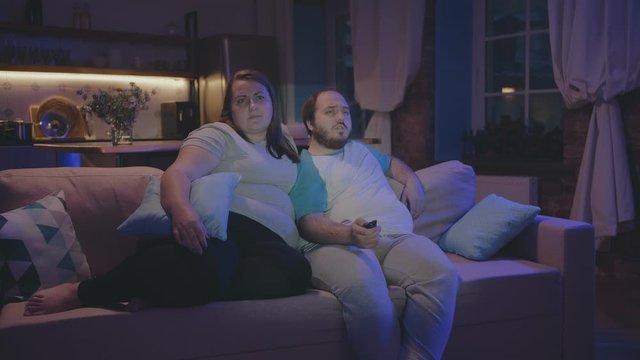 Bored fat couple watching television at home at night