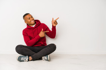 Young latin man sitting on the floor isolated excited pointing with forefingers away.