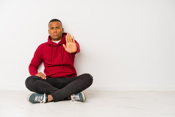 Young latin man sitting on the floor isolated standing with outstretched hand showing stop sign, preventing you.