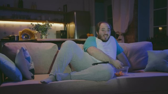 Overweight man lay on the couch with tv remote and junk food.