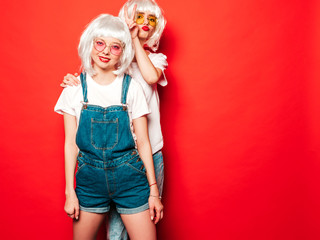 Two young sexy smiling hipster girls in white wigs and red lips.Beautiful trendy women in summer clothes.Carefree models posing near red wall in studio.Positive female going crazy