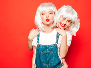 Two young sexy smiling hipster girls in white wigs and red lips.Beautiful trendy women in summer clothes.Carefree models posing near red wall in studio.Positive female going crazy