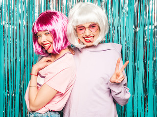 Two young sexy smiling hipster girls in white wigs and red lips.Beautiful trendy women in summer clothes.Carefree models posing on blue silver shiny tinsel background in studio.Positive female