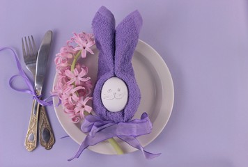 Happy easter concept.Easter holiday table setting with cutlery on light purple background. Pink hyacinth, rubbit from egg and purple napkin with bow on white plate.Top view with copy space for text.