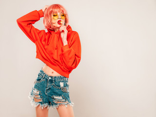 Young beautiful hipster bad girl in trendy red summer red hoodie and earring in her nose.Sexy carefree woman posing in studio on gray background in wig.Hot model licking round sugar candy