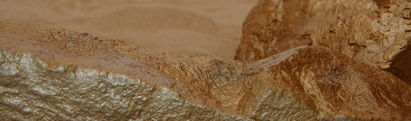A Gold Tone Rock Mineral, Showing the Foreground Top Edge of the Rough Stone, with a Blurred Sand Background.