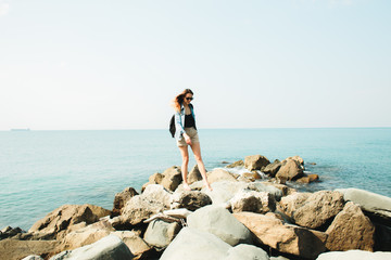a young girl traveler with a backpack walks barefoot on large stones on the sea coast