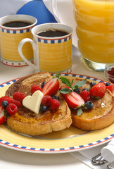 A brunch or breakfast of french toast topped with strawberries, blueberries, and heart shaped butter with coffee and orange juice.