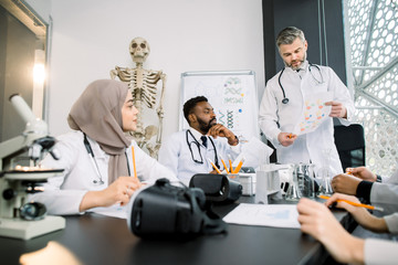 Fototapeta premium Group of multiracial medical students sitting together and studying, listeting to their experienced male professor during the lesson in the classroom. Human skeleton on the background