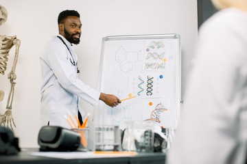 Portrait of young African male doctor or biochemist showing on board some biochemical formulas for his colleagues in conference room. Medicine, biochemistry, science concept